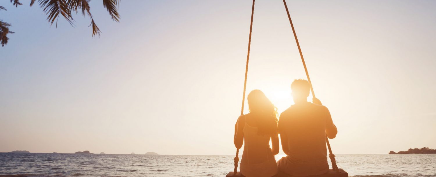 romantic couple in love sitting together on rope swing at sunset beach, silhouettes of young man and woman on holidays or honeymoon