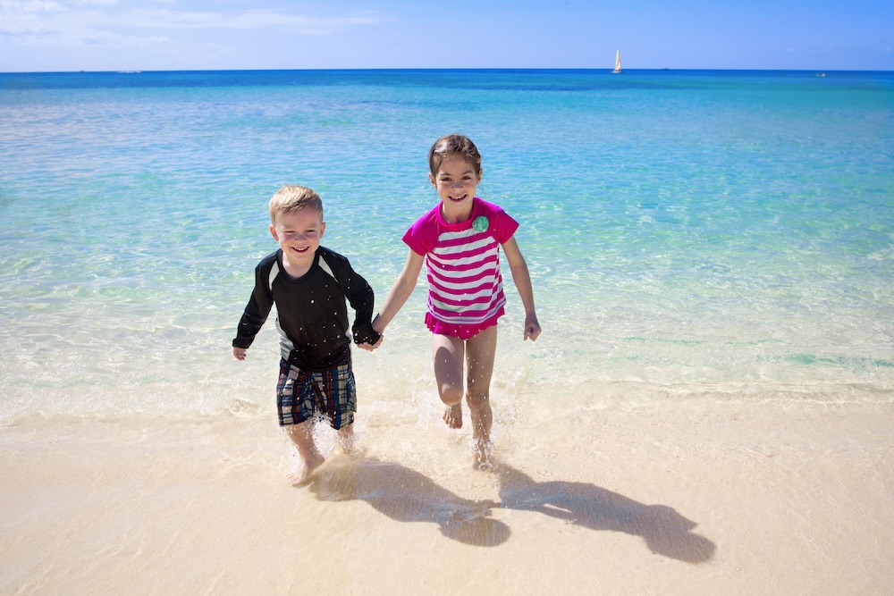 Two cute little kids, a boy and a girl, running along the beach while on a family beach vacation.